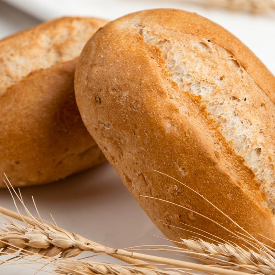 Two rustic golden bread rolls sit on a white plate beside dried wheat stalks.