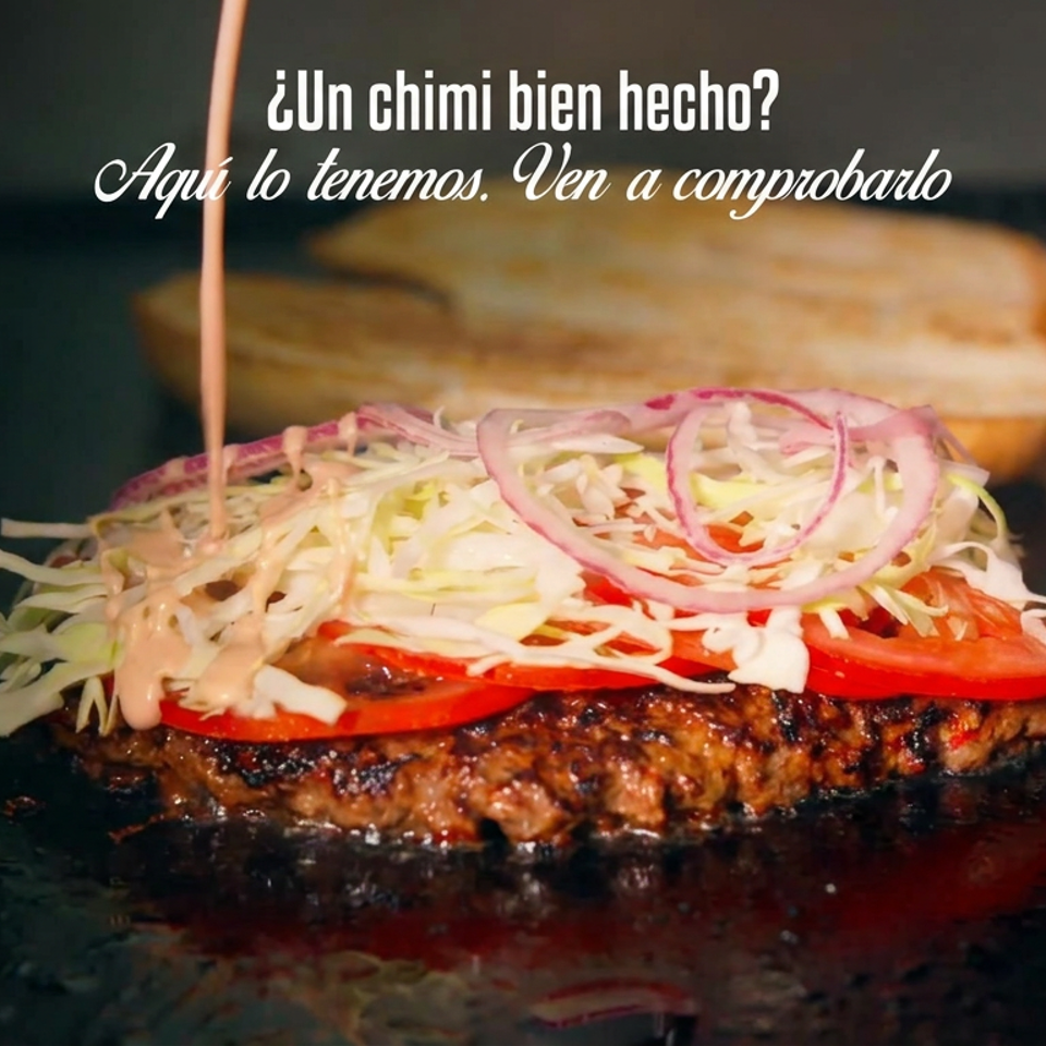 A grilled burger patty is topped with tomato, shredded cabbage, red onion, and sauce on a griddle, with buns behind.
